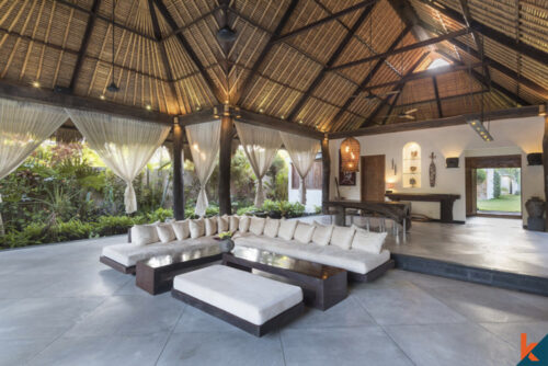 A spacious open-air living area of a Seminyak villa with a high thatched roof, comfortable beige sofas, and sheer drapes, surrounded by lush tropical plants.