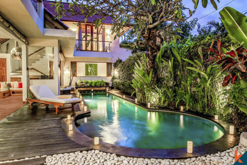 Night view of a serene villa pool in Seminyak with a wooden deck, sun loungers, and vibrant tropical foliage under a starlit sky, reflecting the charm of Balinese outdoor living.