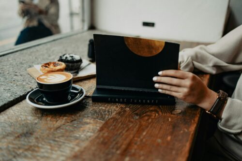 The image captures a moment at a café, with someone's hands holding a black menu with "FINDE ZUKUNFT" in gold, accompanied by a coffee and pastries on a wooden table.