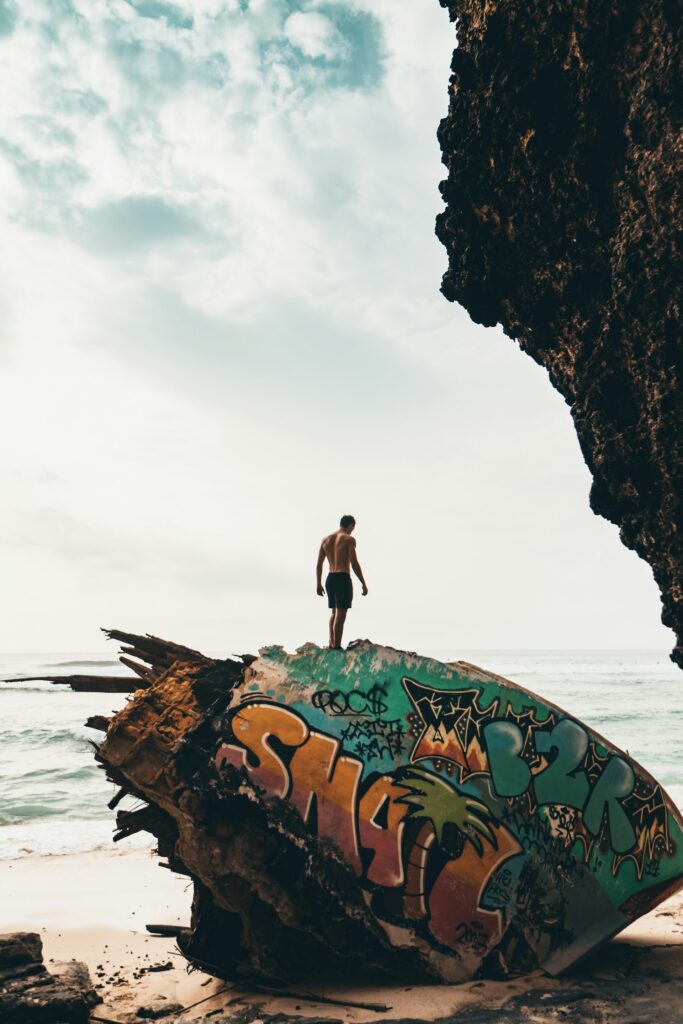 A person standing on a beached surfboard with vibrant graffiti, surrounded by rocks, looking out to the sea.